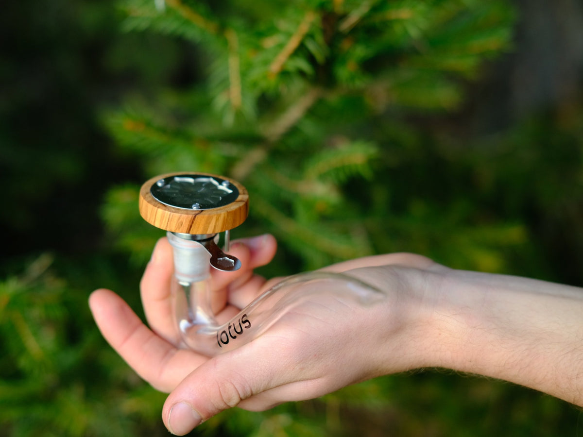 INHALE Lotus Vaporizer mit Glaspfeife in der Hand vor Tannenzweigen